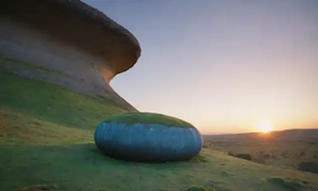 Un jiraft de pie sobre un paisaje de sabanas con un sutil gradiente de tonalidades; su cuerpo alargando de una manera inusual con vibrantes patrones verdes y azules girando; cerca, una entrada cueva oculta con rocas cubiertas de musgo, emitiendo una suave luminiscencia